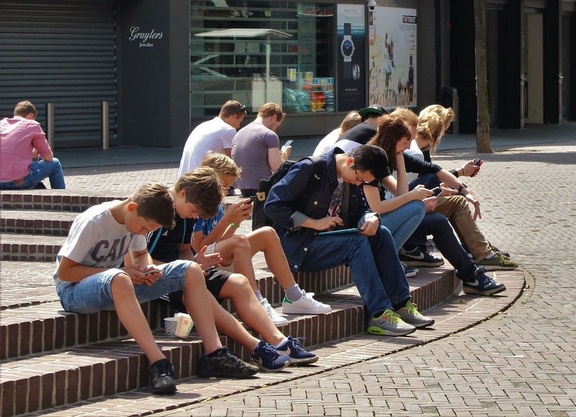 A group of teenagers sitting together on a street corner, all looking down at cell phones or tablets.